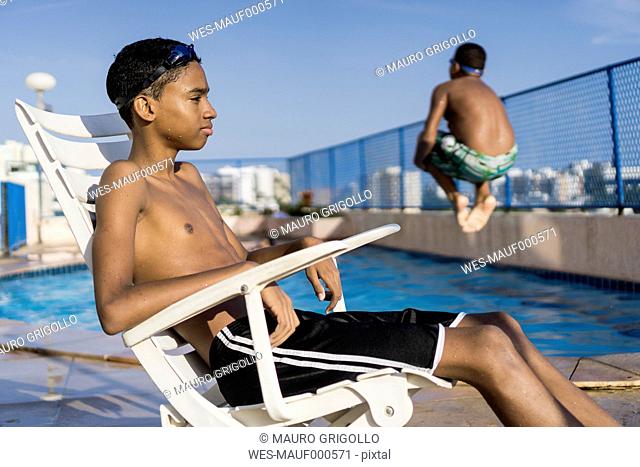 Teenage boy sitting on chair outside swimming pool while his freind is doing a cannonball dive