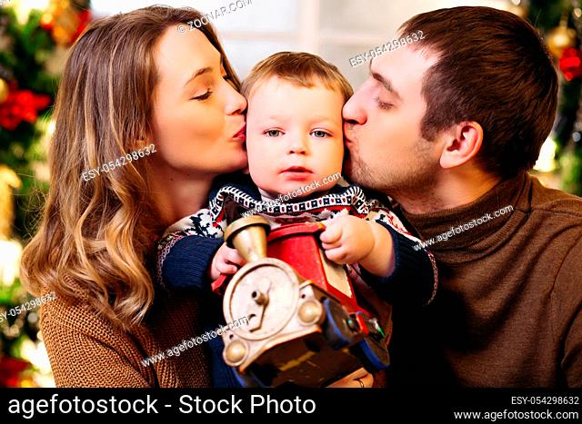 Portrait of loving happy parents kissing their son in cheeks holding toy train as present for Christmas
