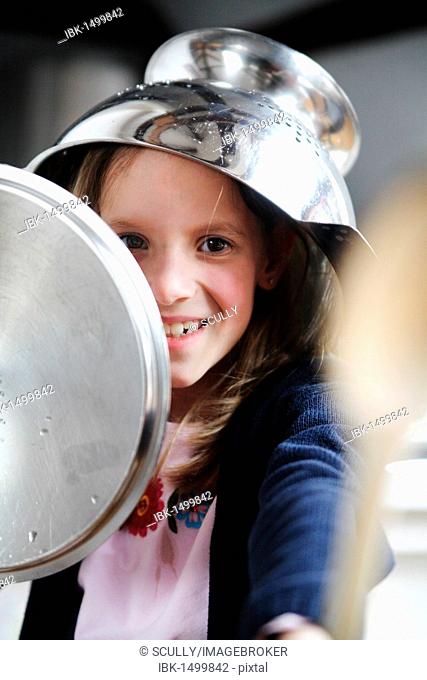 Child in the kitchen with a sieve on her head, spoon in hand and pot lid as a shield