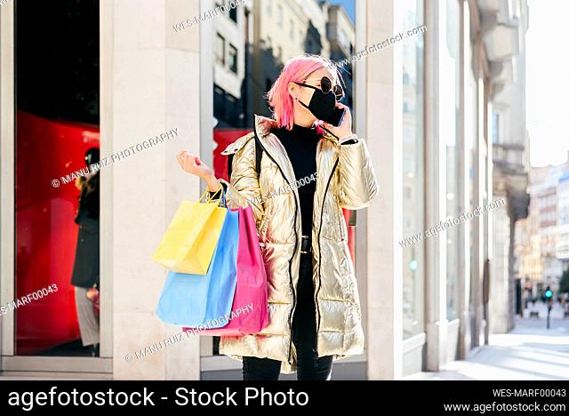Woman wearing protective face mask talking on mobile phone while standing with shopping bags in city