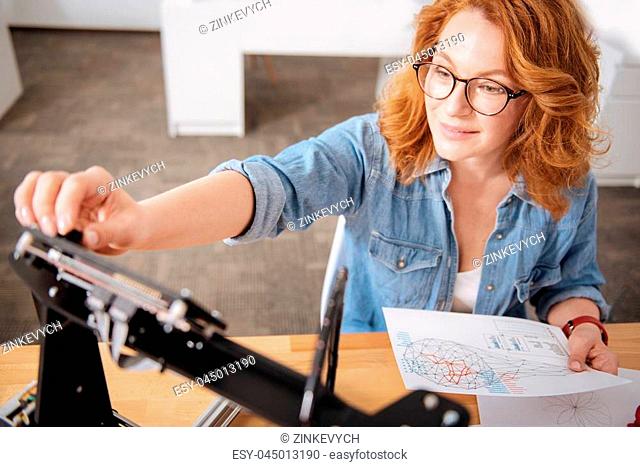 According to the scheme. Nice professional female designer setting up the 3d printer and holding a drawing while preparing to do a 3d graphic design