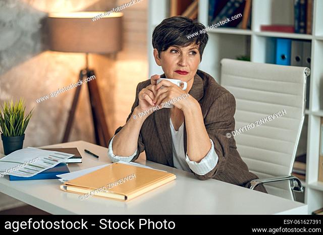 Morning in the office. Good-looking dark-haired woman sitting at the table with a coffee mug in hands