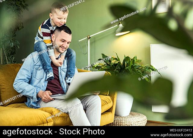 Smiling man using laptop while son sitting on shoulder in living room at home