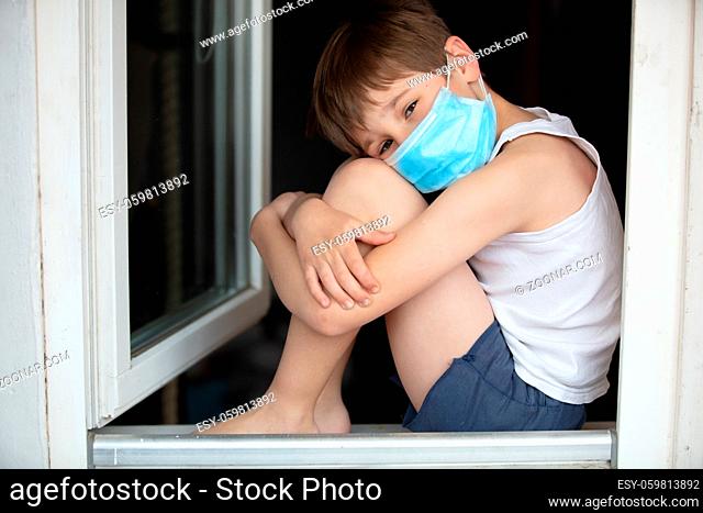 A child in a medical mask sits on a windowsill.Masked quarantine child