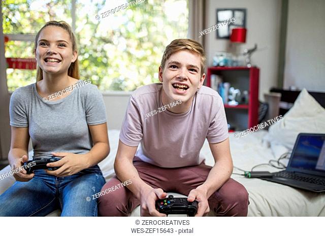 Happy teenage girl and boy sitting on bed playing video game