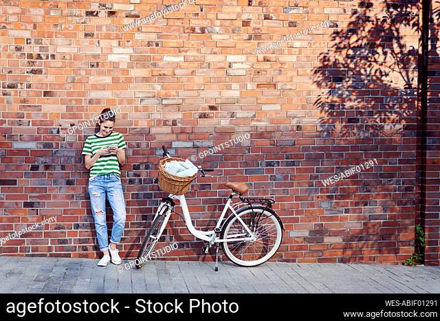 Smiling woman text messaging through smart phone while bicycle parked on footpath against brick wall