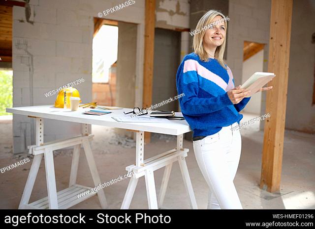 Female building contractor with digital tablet contemplating while leaning on table at apartment