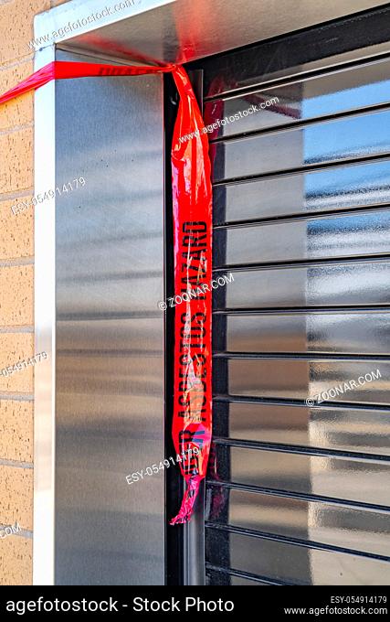 Close up of the window of a building with security roller shutter. A red danger and hazard tape is tied at the corner of the window
