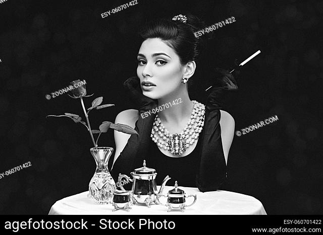 Beautiful young girl looking like audrey hepburn sitting behind table with cigarette. Black and white photo