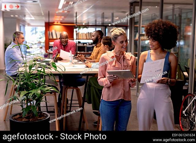 Smiling biracial businesswomen discussing together against colleagues working at modern workplace