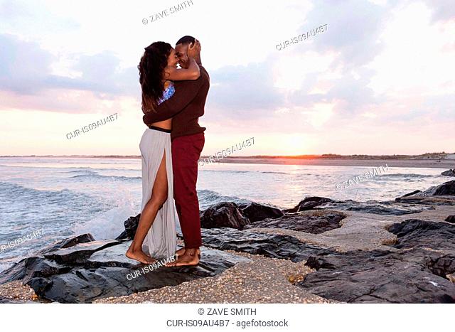 Couple standing on rocks beside sea, hugging, face to face