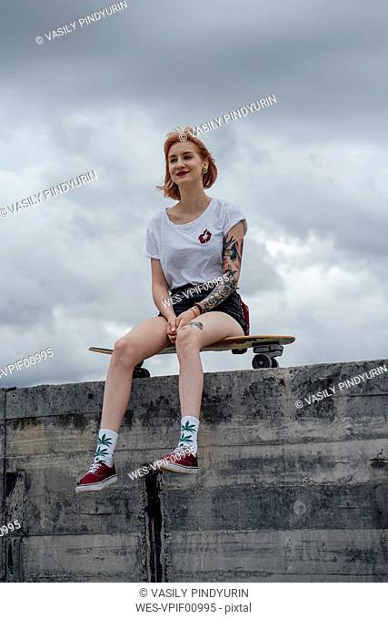 Smiling young woman sitting on a concrete wall on carver skateboard