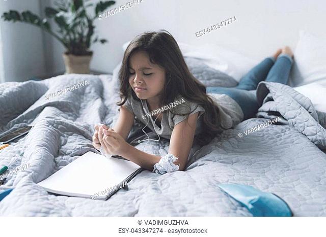 No ideas. A little girl in white headphones is lying on the bed and listening to music next to a white notebook. The girl frowns, since she has no ideas