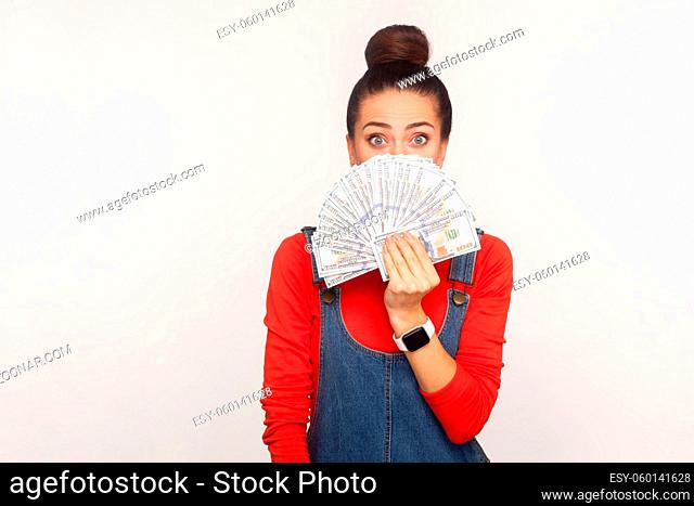 Portrait of astonished girl with hair bun in denim overalls peeking out of money dollars with scared surprised eyes, shocked by lottery win, big profit