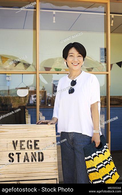 Japanese woman standing outside a farm shop, holding shopping bag, smiling at camera