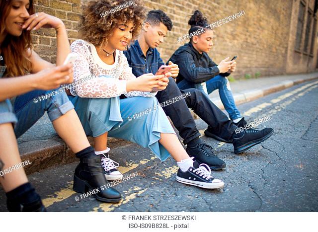 Four friends sitting in street, looking at smartphones