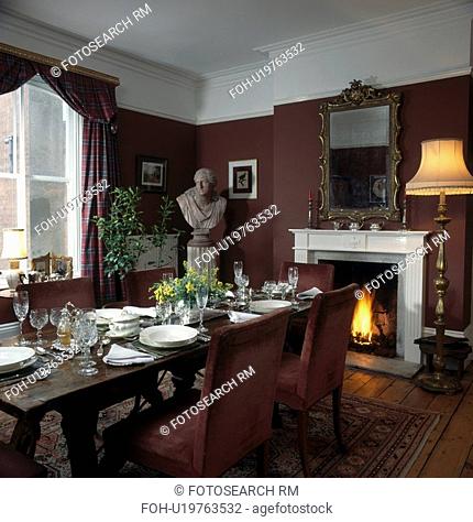 Classical bust in corner of brown dining room with place settings and mirror above lighted fire in fireplace