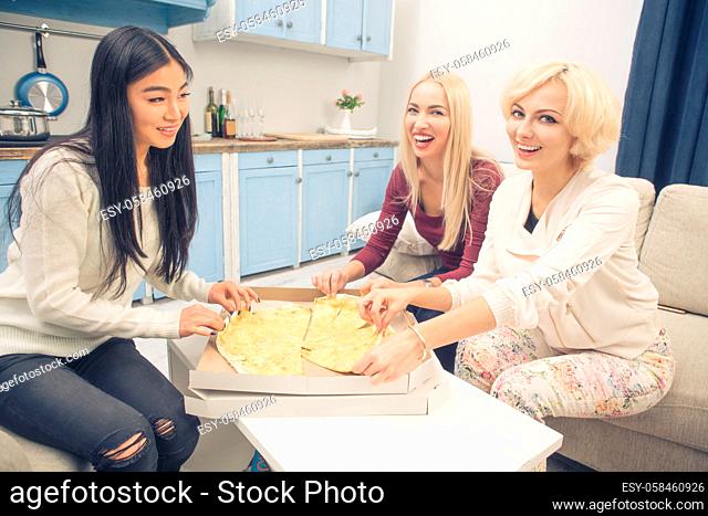 Toned picture of happy best friends girls eating pizza with four cheeses while having party in kitchen at home. Beautiful ladies looking at camera and smiling