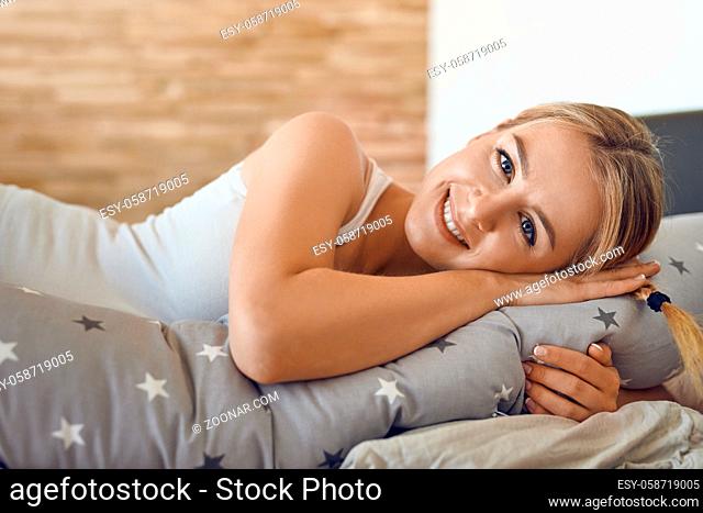 Happy healthy young pregnant woman resting on a bed using a special long pillow as a support looking at the camera with a lovely friendly smile