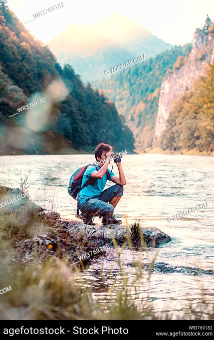 Young tourist with backpack looks through a binoculars on mountains peaks, stands on a rock over a river. Boy spends a vacation in mountains