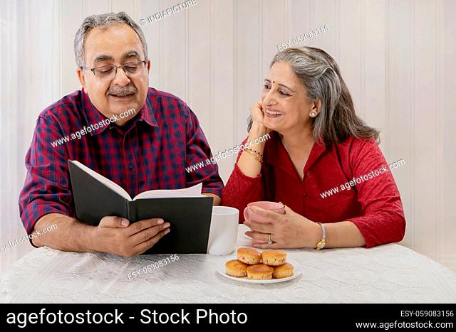 A SENIOR ADULT MAN READING A BOOK WHILE WIFE HAPPILY LISTENS