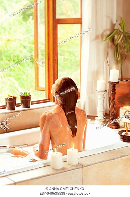 Rear view of a woman sitting in the bath and enjoying water, romantic atmosphere with candles in the bathroom in luxury spa hotel