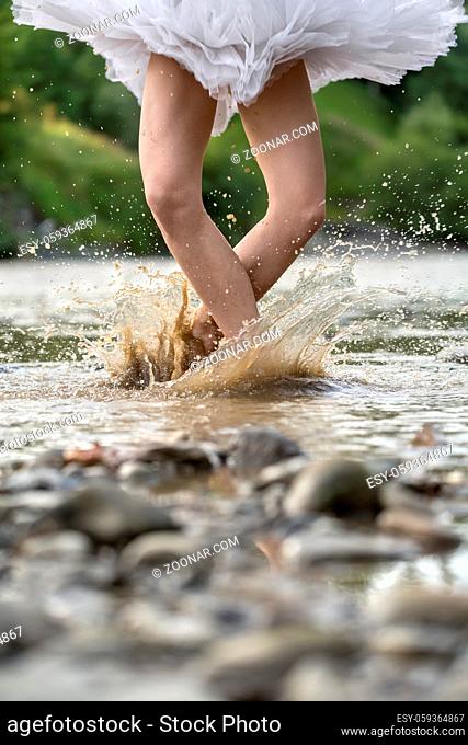 Amazing ballerina jumping in the shallow river on the background of the green shore. She wears a white tutu. Water splashes spreading around her legs