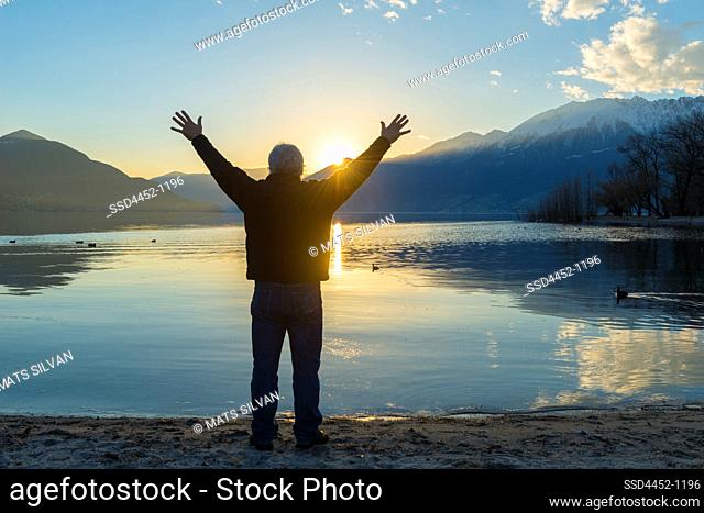 Man with Arms Outstretched Standing on Waterfront on on Alpine Lake Maggiore with Mountain in Sunset in Ascona, Switzerland