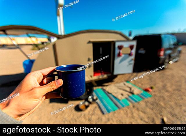 Selective focus of person holding a cup of coffee or tea in front of a parked car and an opened caravan on a camping trip outdoors