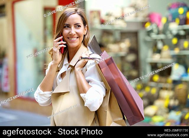 Smiling woman talking on phone while carrying shopping bag in city
