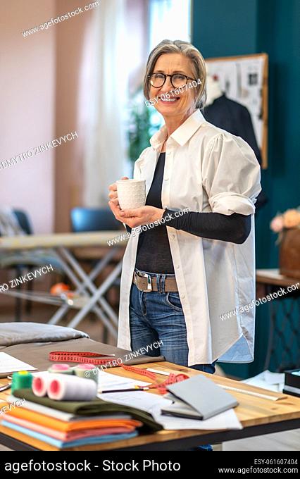 Confidence. Cheerful middle aged woman in glasses with cup standing at workplace in sewing workshop looking confidently at camera