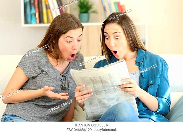 Amazed friends reading newspaper news sitting on a couch in the living room at home
