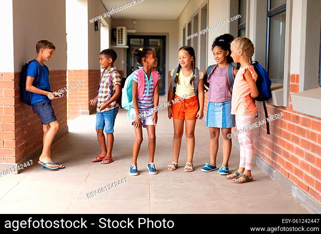 Full length of multiracial elementary school students talking while standing in school corridor