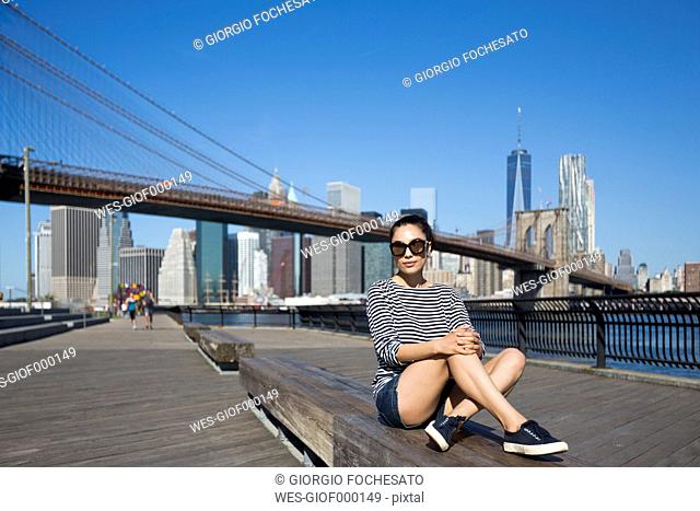 USA, New York City, portrait of young woman relaxing on a bench in front of Brooklyn Bridge