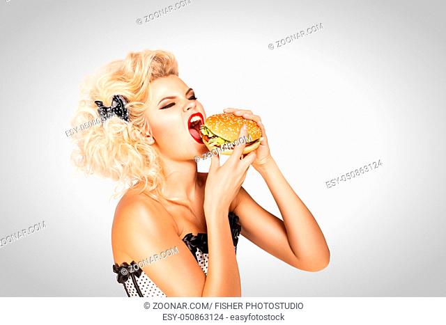 Beautiful pinup model eating a hamburger on grey background