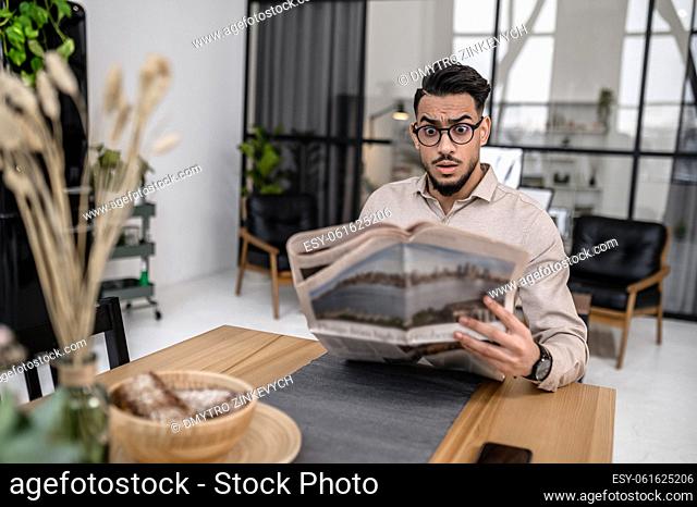 Incredible news. Young bearded good looking man in glasses looking surprised at newspaper while sitting at table in apartment
