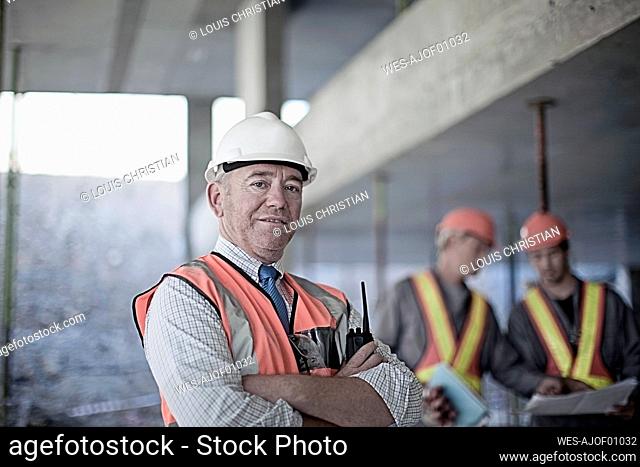 Confident male contractor with arms crossed at site with construction workers seen in background