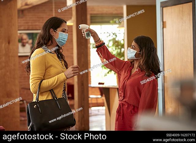 Young ethnic female with purse wearing face mask, having temperature checked with contactless thermometer, by masked co-worker