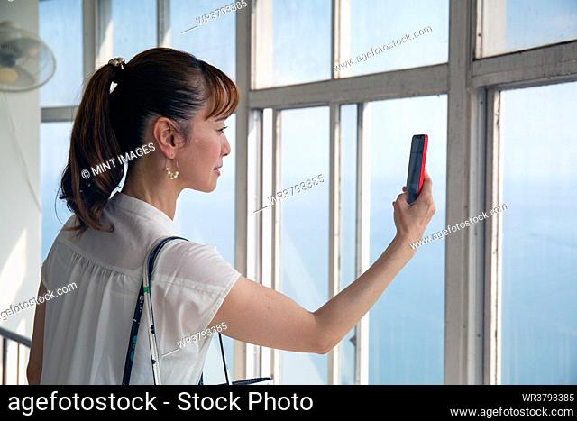 A mature Japanese woman using her mobile phone to take pictures from a viewing platform of the city and landscape below