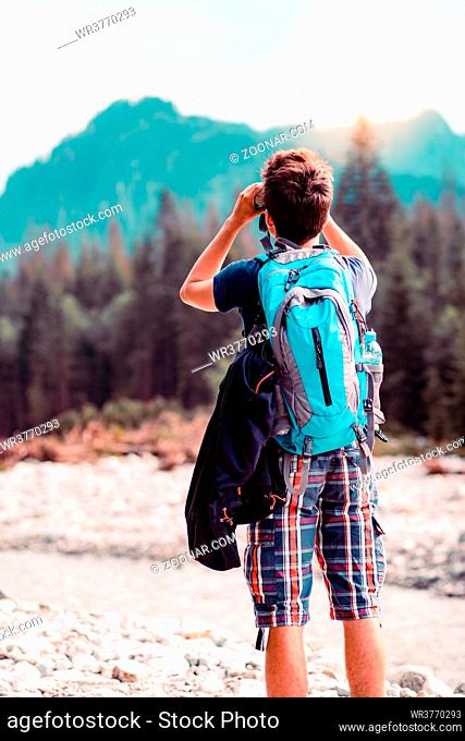 Young wanderer with backpack looks through a binoculars on mountains peaks, stands over a river. Boy spends a vacation in mountains, wandering with backpack