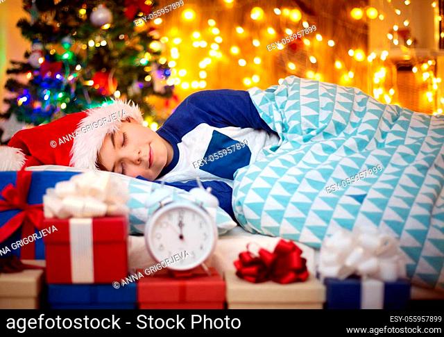 Boy sleeping on the bed with clock near christmas tree. Child in red hat at home in winter