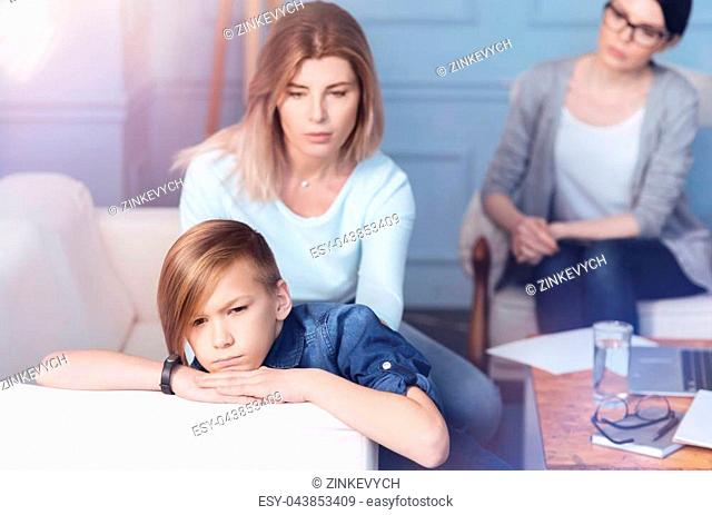 Please look at me. Grumpy male youngster getting irritated while attending a child psychologist with his mom sitting next to him and trying to discuss his...