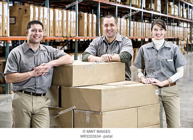 A team portrait of three mixed race uniformed warehouse workers surrounded by boxed products in a large distribution warehouse