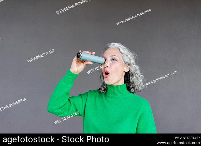 Surprised woman with mouth open looking through monocular in front of wall