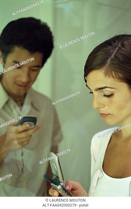 Young woman looking at cell phone, man in background lookng at own phone, smiling