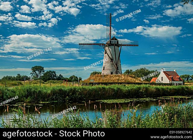Old windmill next to canal with bushes and grove in the background in the late afternoon light and blue sky, near Damme. A quiet and charming countryside old...