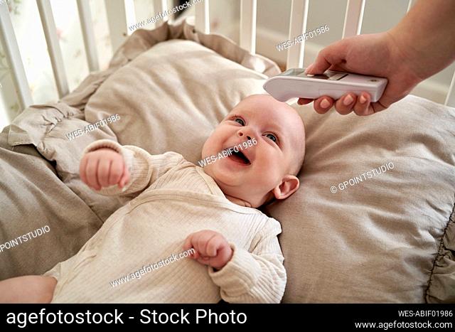 Mother checking temperature of cheerful baby lying on bed