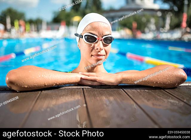 Joyful girl in a white swim cap and swim glasses looks into the camera in the swimming pool outdoors. She holds her hands together on the side of the pool