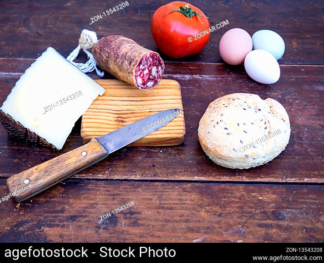 An Iberian pork sausage on an old wooden board with an antique knife, a loaf of Spanish rustic seed bread, cheese, one tomato and three multicolor eggs