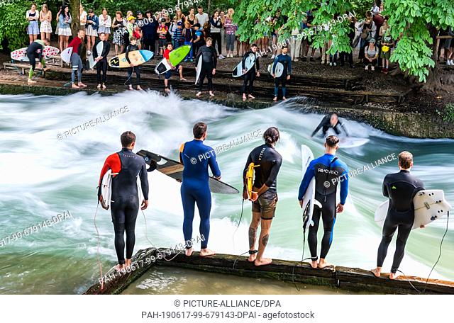 17 June 2019, Bavaria, Munich: Thirteen surfers wait at the edge of the Eisbach wave for their turn. The spot in the English Garden is one of the tourist...